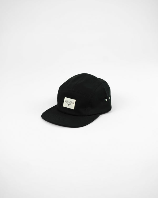 A black five-panel skater cap with an adjustable fastener, flat peak, and no visible brand markings.